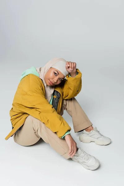Longitud completa de la elegante mujer multirracial en hijab y chaqueta amarilla sentado y mirando a la cámara sobre fondo gris - foto de stock
