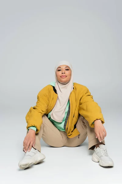 Full length of carefree multiracial woman in hijab and jacket with pants sitting on grey background — Stock Photo