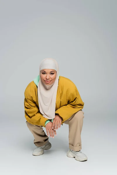 Full length of fashionable multiracial woman in yellow bomber jacket and muslim hijab sitting on haunches on grey background — Stock Photo