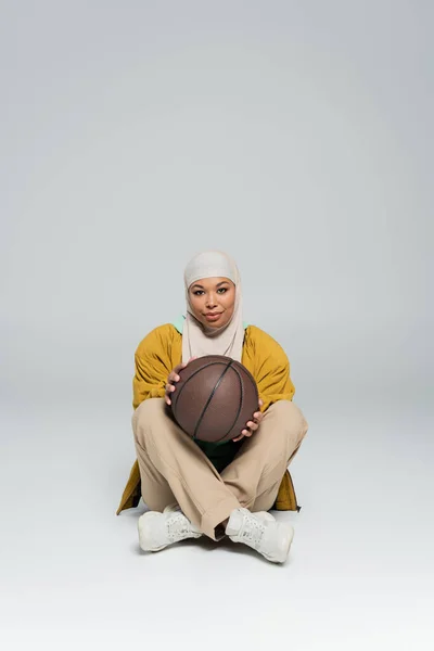 Full length of smiling multiracial woman in hijab and pants with sneakers holding basketball while sitting on grey background — Stock Photo