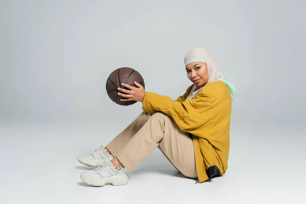 Pleine longueur de femme multiraciale positive en veste de bombardier jaune et hijab tenant le basket tout en étant assis sur fond gris — Photo de stock