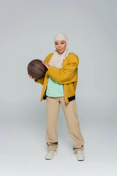 Full length of stylish multiracial muslim woman in yellow bomber jacket and beige pants standing with basketball on grey background — Stock Photo