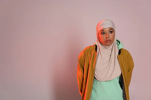 Mujer multirracial reflexiva en chaqueta amarilla y hijab musulmán mirando hacia otro lado sobre fondo gris rosado - foto de stock