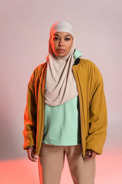 Jovem mulher muçulmana multirracial em casaco de bombista amarelo e hijab olhando para a câmera em fundo cinza e rosa — Fotografia de Stock