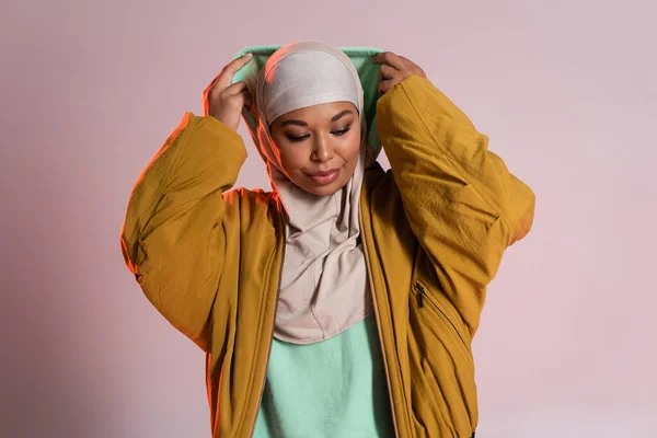 Junge und trendige multirassische Frau in Hijab und gelber Bomberjacke mit Kapuze auf rosa-grauem Hintergrund — Stockfoto