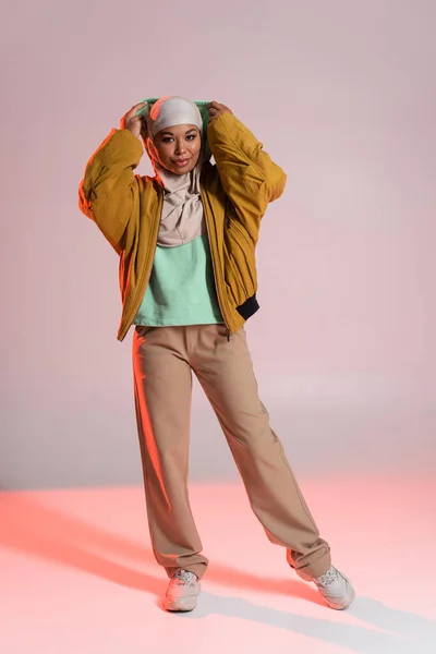 Повна довжина багаторасової жінки в хіджабі і модний повсякденний одяг в капюшоні на сірому і рожевому фоні — стокове фото