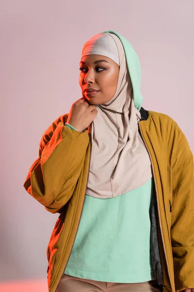 Giovane e bella donna multirazziale in giacca bomber gialla e hijab guardando lontano su sfondo grigio e rosa — Foto stock