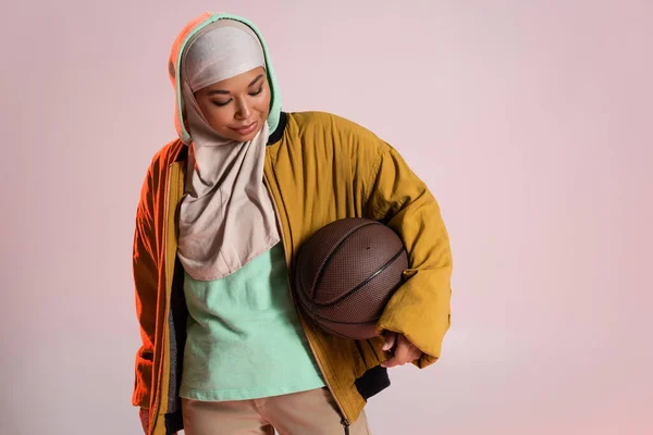 Mujer multirracial de moda en hijab y chaqueta bombardero amarillo que sostiene el baloncesto aislado en gris rosado - foto de stock