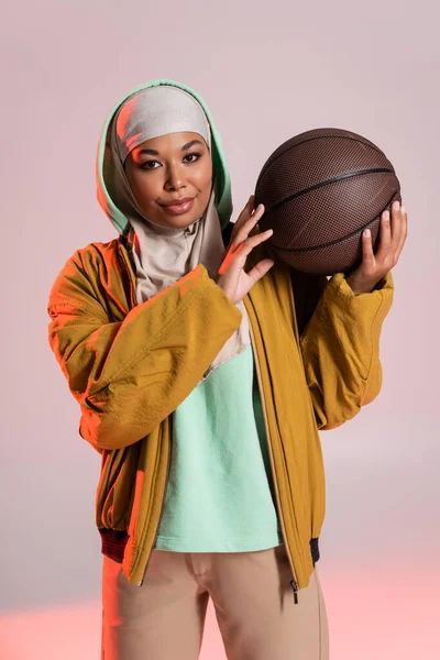 Jolie femme multiraciale en veste jaune et hijab tenant basket tout en souriant à la caméra sur fond gris et rose — Photo de stock