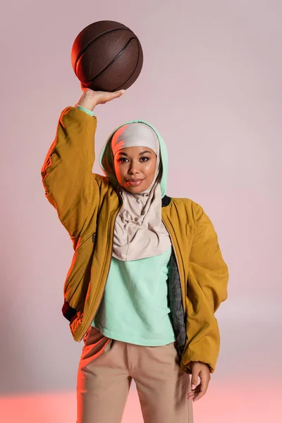 Élégante femme multiraciale en hijab et veste de bombardier jaune debout avec basket dans la main levée sur fond gris et rose — Photo de stock
