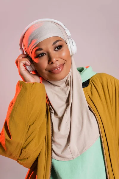 Mujer musulmana multirracial despreocupada en chaqueta amarilla escuchando música en auriculares inalámbricos aislados en gris - foto de stock