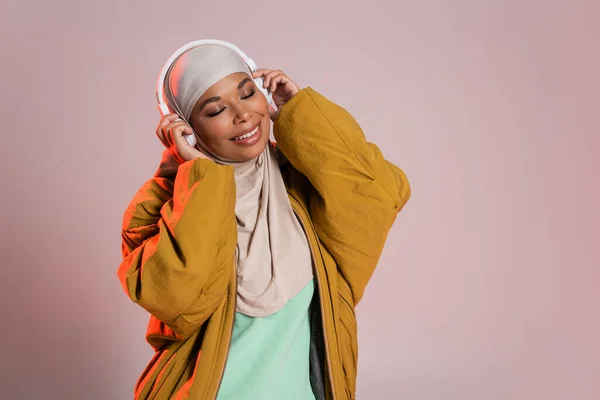 Mujer multirracial llena de alegría en chaqueta de bombardero y hijab escuchando música en auriculares inalámbricos sobre fondo gris rosado - foto de stock