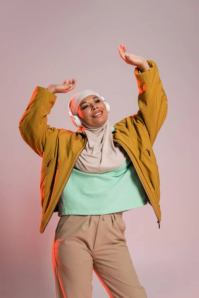 Joyful multiracial muslim woman in stylish casual clothes and headphones dancing with raised hands on pinkish grey background — Stock Photo