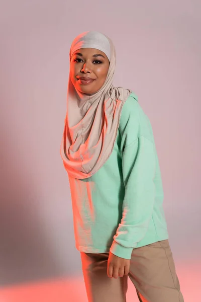 Jolie femme multiraciale en hijab traditionnel et chemise verte à manches longues souriant à la caméra sur fond gris et rose — Photo de stock
