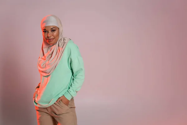 Mulher multirracial sorridente em hijab e camisa de manga longa verde posando com as mãos em bolsos de calças bege no fundo cinza rosado — Fotografia de Stock