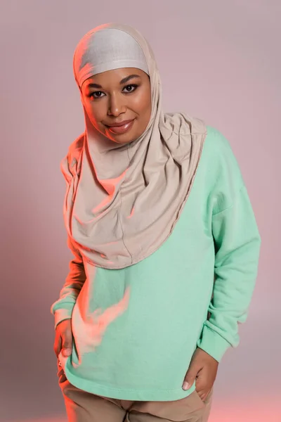 Fashionable multiracial woman in hijab and green long sleeve shirt smiling at camera on pinkish grey background — Stock Photo