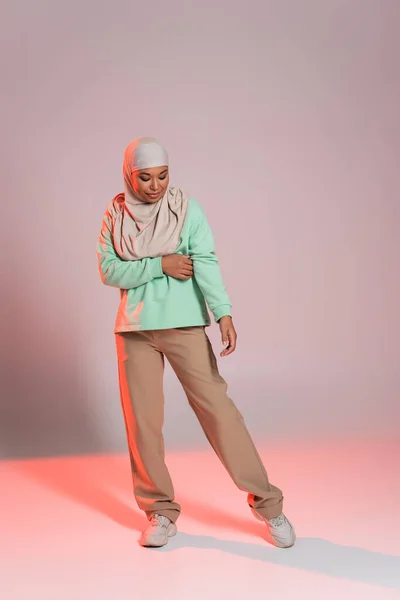 Повна довжина молодої багаторасової жінки в хіджабі і бежевих штанах з кросівками, що стоять на сірому і рожевому фоні — стокове фото