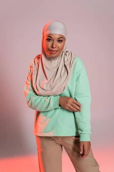 Mulher multirracial bonita em hijab muçulmano e camisa de manga longa verde olhando para a câmera em fundo cinza e rosa — Fotografia de Stock