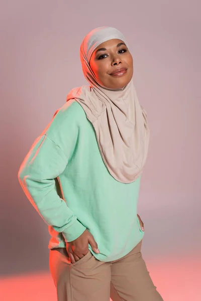 Happy multiracial woman in hijab and green long sleeve shirt posing with hands in pockets on grey and pink background — Stock Photo