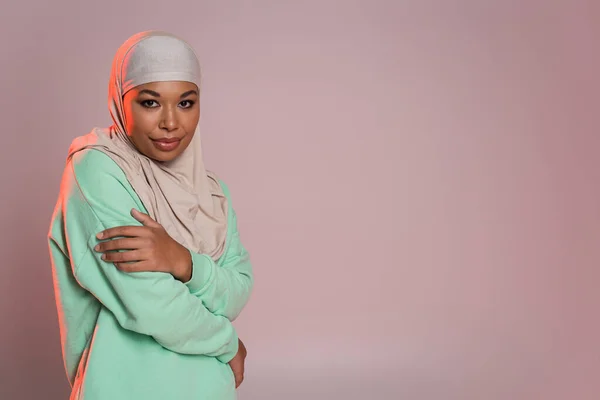 Joven mujer musulmana multirracial en hijab tradicional y camisa verde de manga larga mirando a la cámara mientras posa en gris rosado - foto de stock