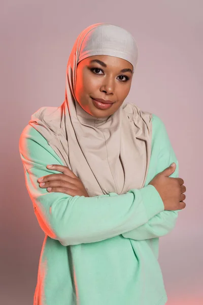 Joven mujer multirracial en hijab y camisa verde de manga larga posando con los brazos cruzados sobre fondo gris rosado — Stock Photo