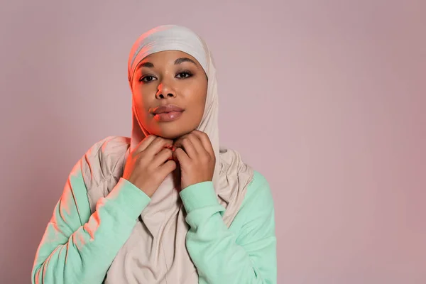 Donna multirazziale positiva in camicia verde a maniche lunghe che regola hijab tradizionale isolato su grigio rosato — Foto stock
