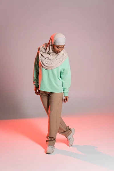 Полная длина многорасовой женщины в хиджабе и рубашке с длинным рукавом с брюками, стоящими на сером и розовом фоне — стоковое фото