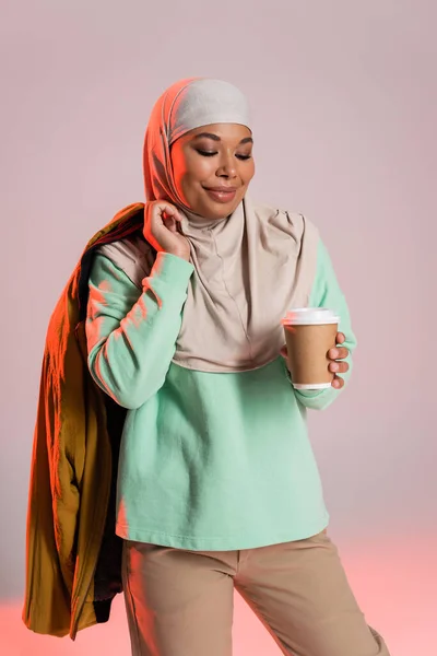 Joyeuse femme multiraciale en hijab tenant veste jaune et boisson à emporter dans une tasse jetable sur fond gris et rose — Photo de stock