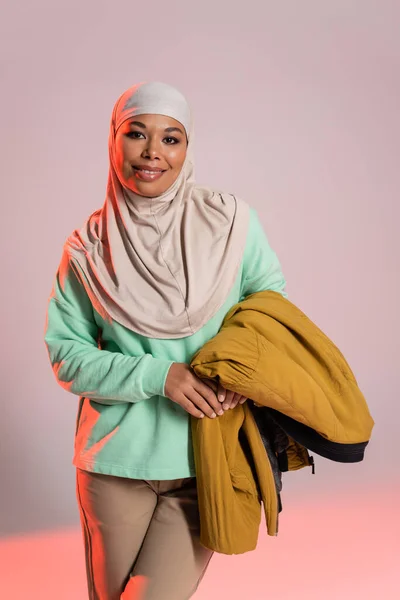 Cheerful multiracial muslim woman in hijab holding yellow jacket and smiling at camera on grey and pink background — Stock Photo