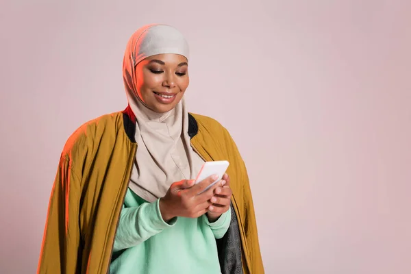 Femme multiraciale heureuse en veste de bombardier jaune et hijab à l'aide d'un téléphone mobile isolé sur gris rosé — Photo de stock