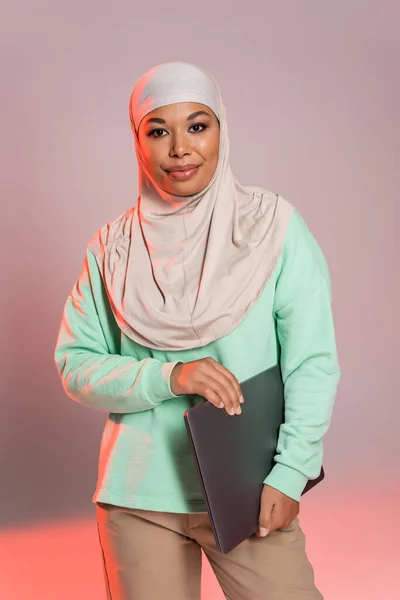 Mujer multirracial feliz en hijab sosteniendo el ordenador portátil y sonriendo a la cámara sobre fondo gris y rosa - foto de stock