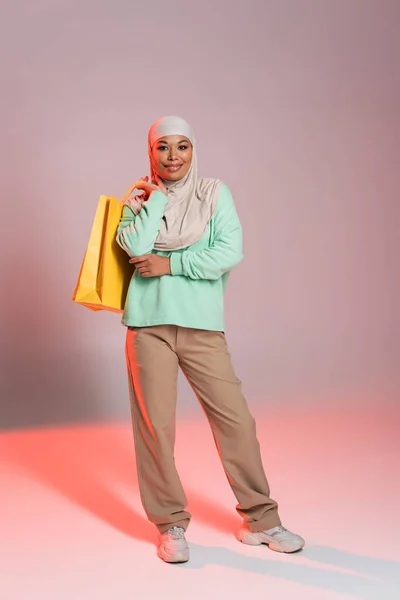 Pleine longueur de femme multiraciale heureuse en hijab et tenue décontractée tendance posant avec des sacs à provisions jaunes sur fond gris et rose — Photo de stock