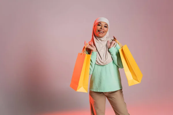 Радостная многорасовая женщина в модном повседневном одеянии и традиционном хиджабе, держащая желтые пакеты для покупок на розово-сером фоне — стоковое фото
