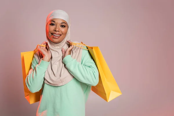 Femme multiraciale ravie en hijab musulman tenant des sacs à provisions jaunes et souriant à la caméra sur gris rosé — Photo de stock