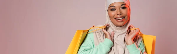 Беззаботная многорасовая женщина в хиджабе позирует с желтыми сумками для покупок и улыбается в камеру, изолированную на сером, баннер — стоковое фото