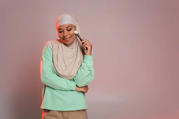 Mujer musulmana multirracial complacida en hijab sosteniendo cepillo cosmético y sonriendo con los ojos cerrados sobre fondo gris rosado - foto de stock
