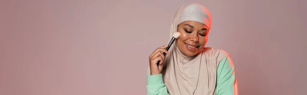 Sorrindo mulher multirracial no hijab segurando escova cosmética enquanto de pé com os olhos fechados isolados em cinza rosado, banner — Fotografia de Stock