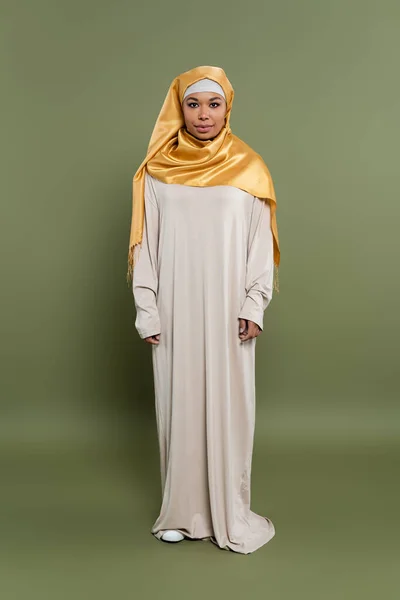 Pleine longueur de femme musulmane multiraciale en hijab de soie jaune et robe abaya debout sur fond vert olive — Photo de stock
