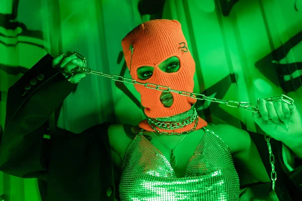 Провокаційна жінка в в'язаному Балаклаві і срібному верхньому тримає металевий ланцюг біля стіни з графіті в зеленому освітленні — стокове фото