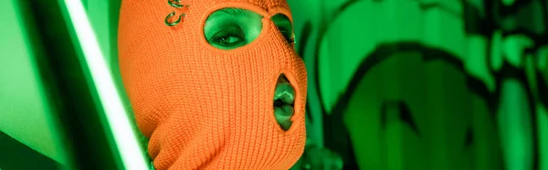 Портрет сексуальної жінки в в'язаній помаранчевій балаклаві біля яскравої неонової лампи і зеленої стіни з графіті, банер — стокове фото