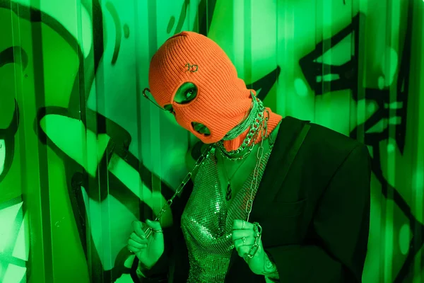 Seducente donna in passamontagna arancione e blazer nero con top in argento in posa con catena metallica vicino al muro verde con graffiti — Foto stock