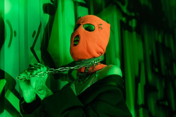 Татуированная женщина в оранжевой балаклаве держит металлическую цепь на шее возле стены с граффити в зеленом свете — стоковое фото
