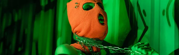 Femme séduisante en cagoule orange tenant des chaînes de cou métalliques près du mur vert avec graffiti, bannière — Photo de stock