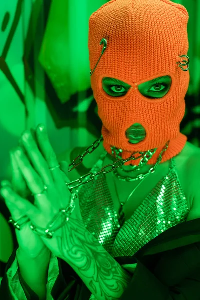 Provocative woman in orange balaclava and metallic top with neck chains looking at camera near wall with graffiti in green light — Stock Photo