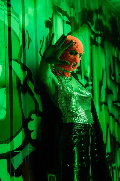 Femme sexy en cagoule et haut en argent debout avec la main tendue près du mur avec des graffitis dans la lumière verte néon — Photo de stock