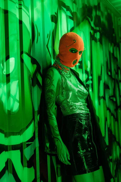 Сексуальна жінка в чорній шкіряній спідниці та срібному верху з апельсиновою балаклавою, що стоїть біля зеленої стіни з графіті — стокове фото