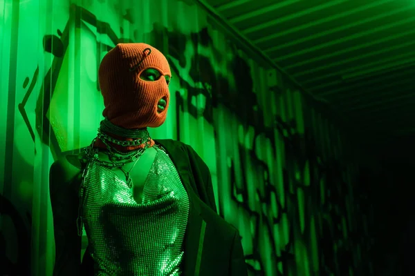 Анонимная сексуальная женщина в оранжевой балаклаве и серебряном топе, смотрящая на стену с граффити в зеленом свете — стоковое фото
