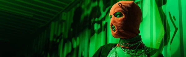 Femme anonyme sexy en balaclava orange et colliers argentés regardant loin dans la lumière verte près du mur avec graffiti, bannière — Photo de stock
