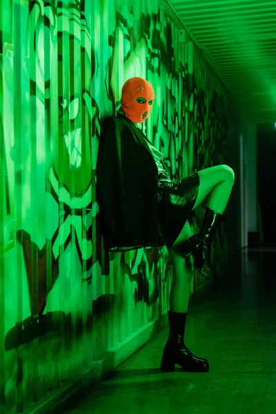 Full length of sexy woman in stylish leather boots and orange balaclava standing near wall with graffiti in green lighting — Stock Photo