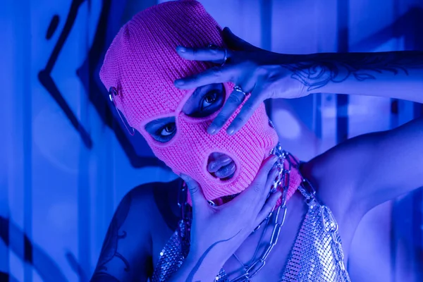 Ritratto di seducente donna in passamontagna e top argentato in posa con le mani vicine faccia a parete con graffiti in luce blu al neon — Foto stock